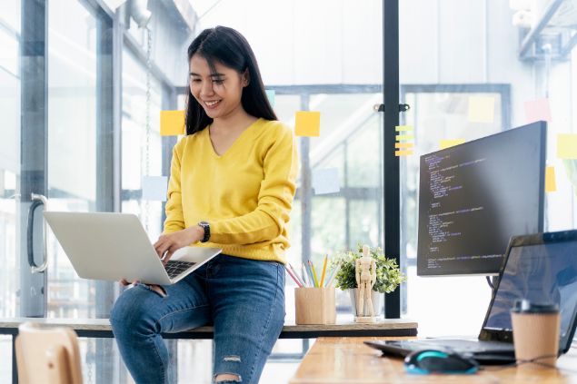 Imagem de uma mulher sentada com seu notebook em cima da mesa do seu escritório, sorrindo