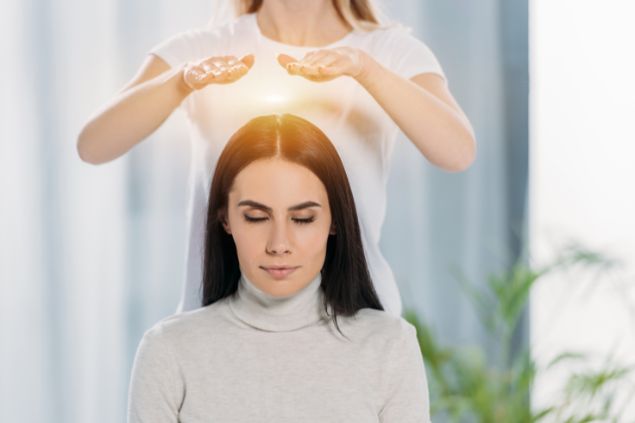 LightFieldStudios / Getty Images / Canva Imagem de uma mulher sentada, recebendo a aplicação do reiki pelas mãos de uma reikiana.