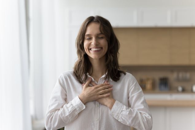 Fizkes / Getty Images / Canva Imagem de uma mulher usando uma camisa branca, ela está sorrindo e feliz, com os olhos fechados, com as mãos sobre o peito, simbolizando o gesto de gratidão.