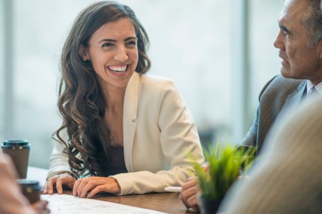 Imagem de uma mulher de cabelos longos sorrindo em seu escritório de trabalho. Ao lado dela, está um homem usando terno e gravata.