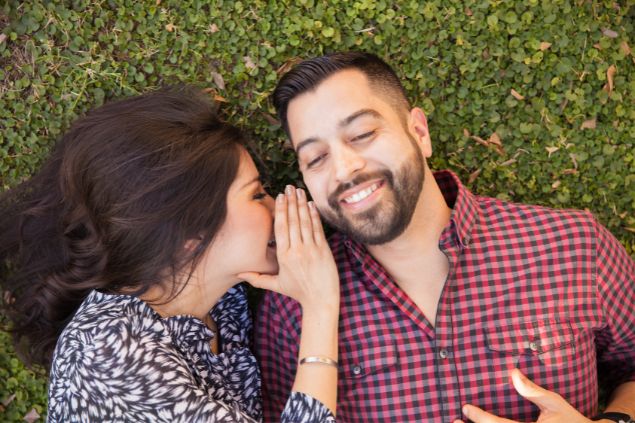 Imagem de um casal jovem, deitado sobre uma grama. Ela está falando ao ouvido dele e ambos sorriem.