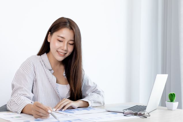 Imagem de uma mulher de cabelos longos, usando uma camisa de listras clara. Ela está em sua mesa, analisando algumas planilhas, no escritório do trabalho.